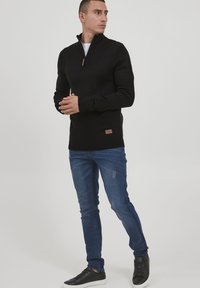 Sweater noir zippé au cou avec des poignets et un ourlet côtelés, porté sur une chemise blanche. Associé à un jean bleu et des baskets noires avec une semelle blanche.