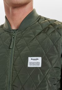 Zelená prošívaná bomber bunda se zipem vpředu, žebrovaným límcem a štítkem s textem. Materiál má hladký, lesklý povrch.