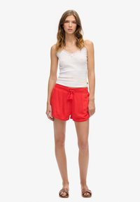 Pantalones cortos rojos con un patrón texturizado y una cintura con cordón, combinados con una camiseta sin mangas blanca con detalles de encaje. Las sandalias completan el conjunto.