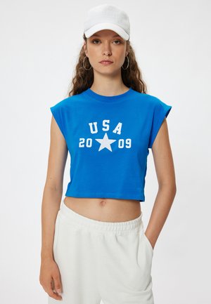 Camiseta corta azul con mangas enrolladas, con texto blanco "USA 2009" y un gráfico de estrella. Combinada con pantalones blancos de cintura elástica.