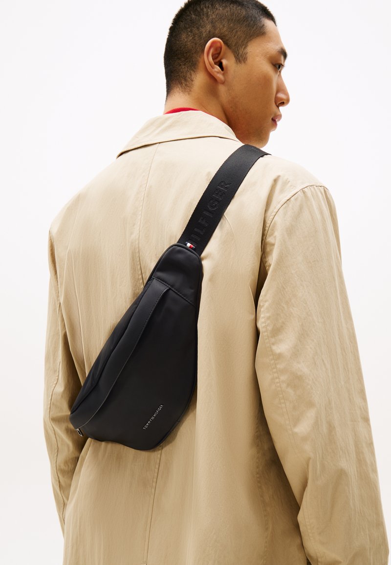 Sac crossbody noir en nylon lisse avec une forme incurvée. Il dispose d'une sangle texturée avec une marque et d'une petite touche de couleur sur la quincaillerie.