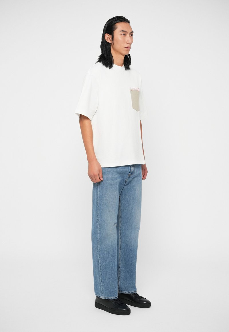 Weißes Baumwoll-T-Shirt mit oversized Schnitt und Tasche auf der linken Brust. Kombiniert mit hellblauen Jeans und schwarzen Turnschuhen.