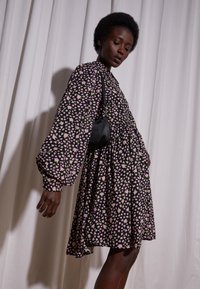 Vestido negro com mangas bufantes longas, apresentando um padrão floral em rosa e verde. O tecido tem uma textura suave e cai ligeiramente acima do joelho.