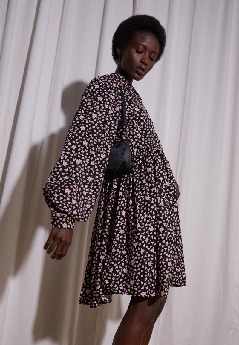 Vestido negro com mangas bufantes longas, apresentando um padrão floral em rosa e verde. O tecido tem uma textura suave e cai ligeiramente acima do joelho.