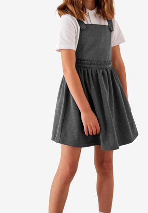 Dziecko ubrane w ciemnoszarą sukienkę na szelkach z guzikami, na białej koszuli z krótkim rękawem i kołnierzykiem, stojące na białym tle.