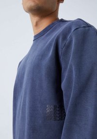 Sudadera azul marino de tejido suave con un cuello redondo y un corte ligeramente oversize. Presenta un logo texturizado en la manga izquierda.