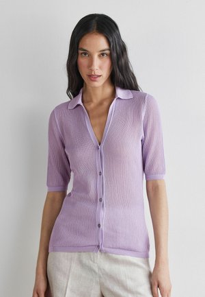 Frau mit langen dunklen Haaren, die eine hellviolette kurzärmelige, geknöpfte Strickjacke und beige Hosen trägt, steht vor einer schlichten weißen Wand.