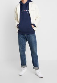 Man som bär en marinblå hoodie med vitt tryck, beige jacka, uppvikt blå jeans och vita sneakers, står med händerna i fickorna.