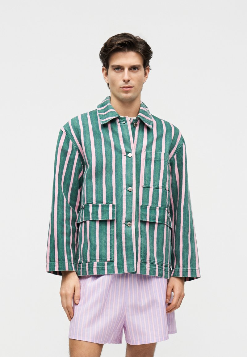 Bărbat purtând o jachetă cu dungi verticale verzi și roz, cu buzunare frontale și nasturi, asortată cu pantaloni scurți cu dungi mov deschis și roz, stând nemișcat.