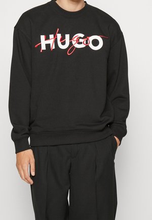 Osoba nosząca czarną bluzę z kapturem z napisem "HUGO" w białych i czerwonych stylizowanych literach na piersi, zestawioną z czarnymi spodniami.