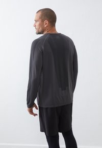 Langarm sportshirt in donkergrijs met een meshstructuur, voorzien van een ronde hals en een contrasterend patroon op de rug. Gecombineerd met zwarte shorts.