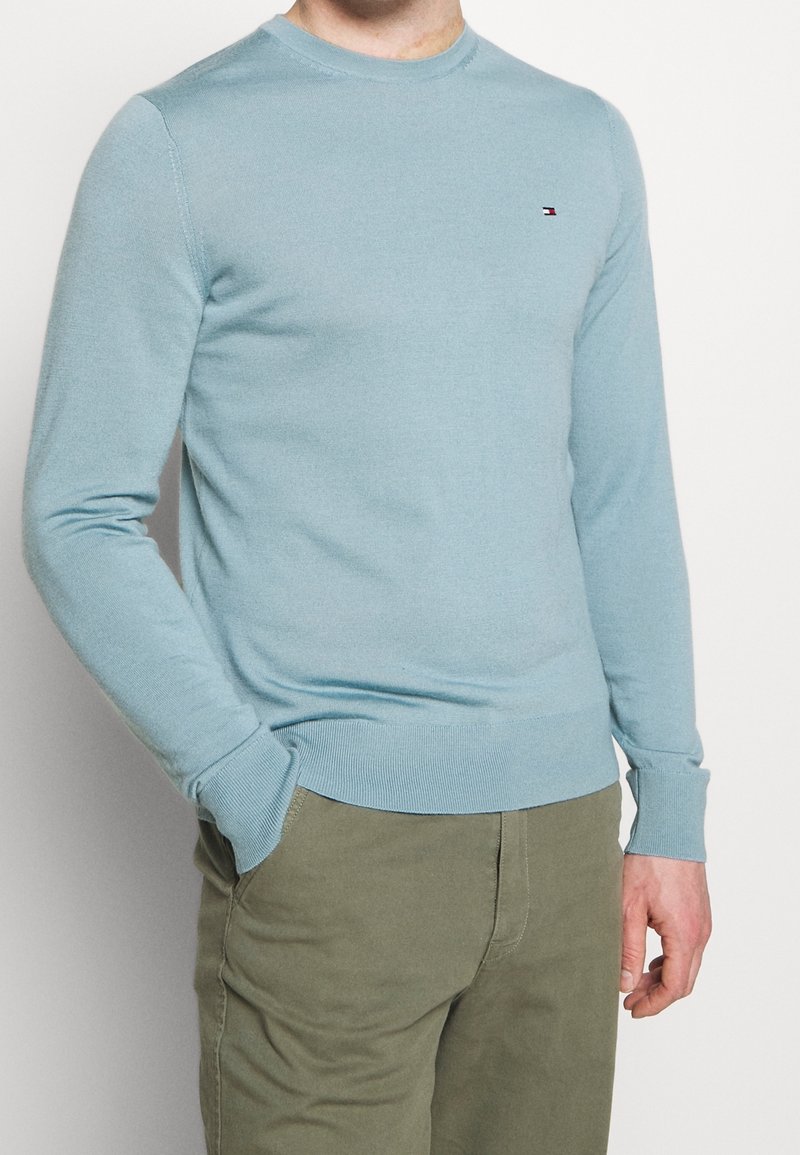 Homme portant un pull léger bleu clair à manches longues avec un petit logo sur la poitrine et un pantalon vert olive, debout avec une main dans la poche.