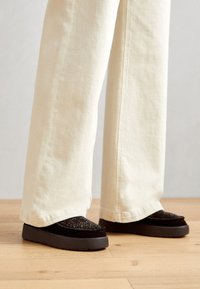 Zapatos negros tipo slip-on con parte superior texturizada, adornados con pequeñas tachuelas y suela gruesa de goma gris. Combinados con pantalones de pierna ancha color crema.