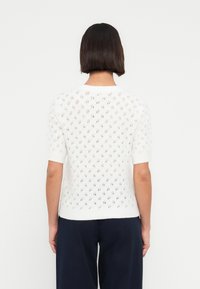 Λευκό πλεκτό πουλόβερ με κοντά μανίκια, ανοιχτό πλέγμα σε σχήμα διαμαντιού και στρογγυλή λαιμόκοψη. Το κάτω μέρος έχει ριμπ τελείωμα.