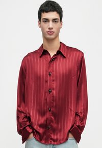 Young man wearing a shiny red striped button-up shirt with decorative buttons, hands in pockets, against a plain white background.