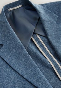 Detailní záběr na modrý blazer s texturou, ukazující límec, klopu a hladkou tmavě modrou podšívku s kontrastním světlým lemováním.