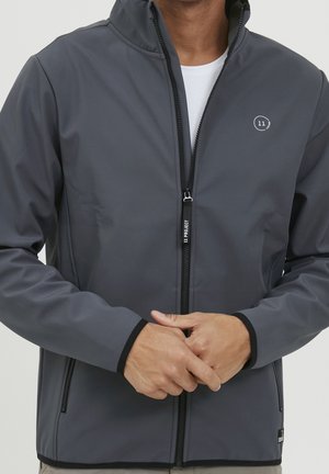 Homme portant une veste zippée gris foncé avec bordure noire, une chemise intérieure blanche et un logo circulaire "11 Project" sur la poitrine gauche.