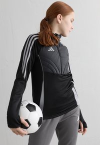 Melnas sporta jakas ar rāvējslēdzēja apkakli, kvadrātu augšējo paneli un pelēkajiem akcentiem. Modelis tur melnbaltu futbolbumbu, valkājot pelēkas bikses.