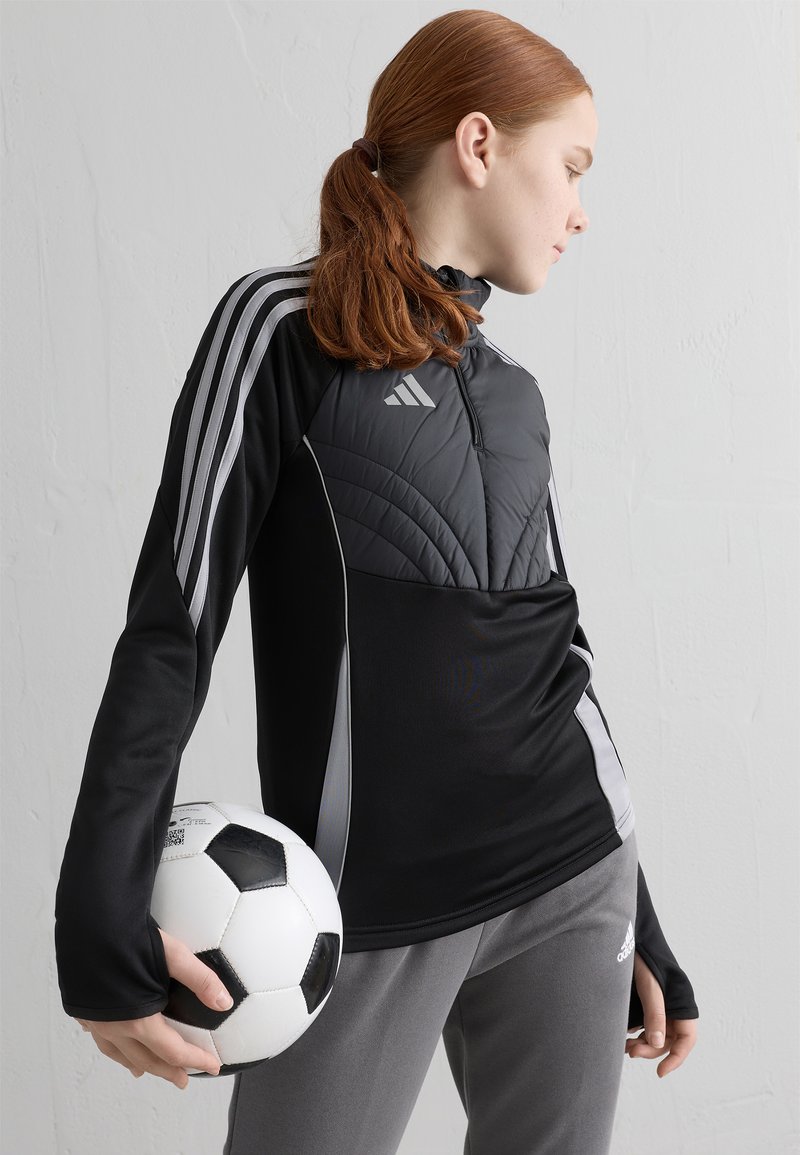 Melnas sporta jakas ar rāvējslēdzēja apkakli, kvadrātu augšējo paneli un pelēkajiem akcentiem. Modelis tur melnbaltu futbolbumbu, valkājot pelēkas bikses.