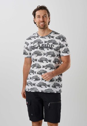 Mann trägt weißes T-Shirt mit schwarzen Fischgräten-Prints und schwarze Shorts, steht vor einem einfarbigen hellen Hintergrund und lächelt.