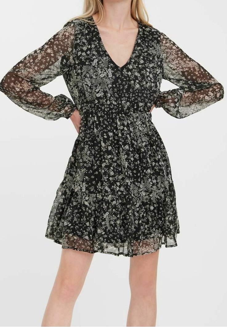 Robe noire à fleurs avec un col en V, des manches longues en voile transparent et une jupe à volants. Le tissu présente un motif floral délicat dans des teintes claires.