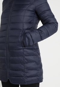 Marinblå pufferjacka med vertikala quiltade mönster, dragkedjestängning och sidofickor. Slätt, glänsande tyg med rak silhuett och rundad nederkant.