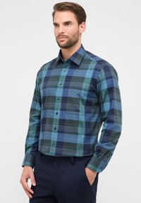 Uomo che indossa una camicia a maniche lunghe a quadri blu e verdi e pantaloni scuri, in piedi con una mano in tasca su uno sfondo semplice.