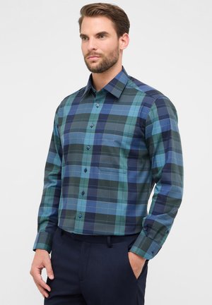 Man in een blauw en groen geblokt overhemd met lange mouwen en donkere broek, staande met één hand in zijn zak tegen een effen achtergrond.