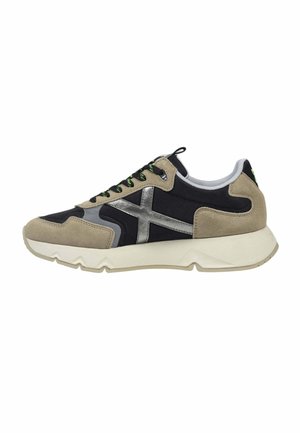 Zapatilla baja con ante beige y paneles de tela negra, logo lateral plateado, suela blanca y cordones negros con detalles verdes, vista lateral.