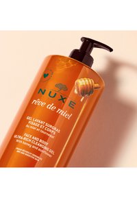 Pomarańczowa plastikowa butelka z pompką, z ilustracją miodu, zielonym tekstem i białą etykietą, na której szczegółowo opisano typ produktu i składniki.