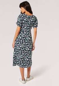 Blumenkleid mit Midi-Länge, navy Hintergrund, weißem und grünem Blumenmuster, kurzen Puffärmeln und einer lockeren Passform. Dazu weiße Schuhe.