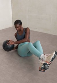 Abbigliamento sportivo composto da una canotta nera, leggings verde menta e scarpe beige con dettagli neri. Tiene in mano una palla medica nera.