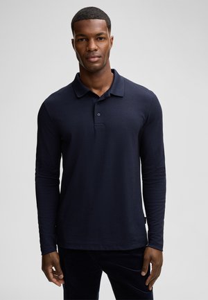 A man with short hair and a serious expression wears a navy long-sleeve polo shirt, standing against a light gray background.