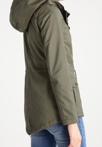 Widok z boku osoby noszącej oliwkowozieloną kurtkę z kapturem z zapinanymi na guziki kieszeniami oraz niebieskie dżinsy.