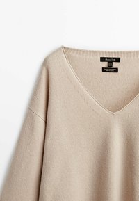 Beige wollen-kashmir trui met een V-hals, ribbelrand en zachte textuur. Labels geven materiaal- en verzorgingsinstructies aan.