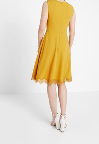 Žluté šaty do poloviny stehna s vybroušeným korzetem a rozšířenou sukní. Krajkový lem s vlnkami zdobí spodní okraj a má jemný detail na zadním švu.
