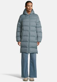 Eine Frau trägt einen langen grauen, gepolsterten Kapuzenmantel, weit geschnittene blaue Jeans und beige Schuhe und steht vor einem schlichten Hintergrund.