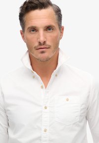 Camisa blanca de botonadura con cuello de punta, hecha de algodón, con un bolsillo en el lado izquierdo y botones beige en el frente.