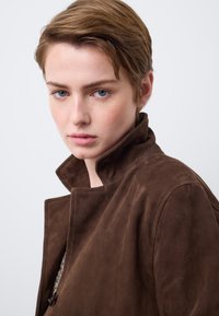 Braune Wildlederjacke mit Stehkragen, die eine weiche Textur und klare Linien aufweist, teilweise sichtbar über einem hellfarbenen Oberteil.