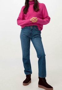 Pull à col roulé côtelé rose, jean taille haute bleu, et baskets compensées marron avec lacets. Accent sur la texture et la coupe moderne.