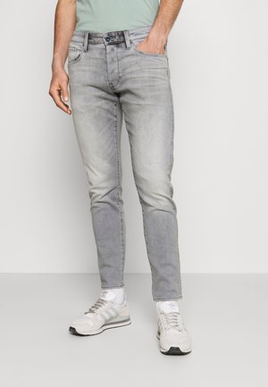Man draagt lichtgrijze slim-fit jeans, witte sneakers en een lichtgroen shirt, staand met één hand in de zak.