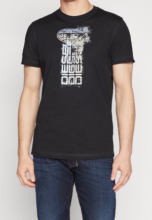 Mann trägt ein schwarzes kurzärmliges T-Shirt mit vertikalem distressed Grafikmotiv und verblasstem blauem Kreis-Design, kombiniert mit dunkelblauen Jeans.