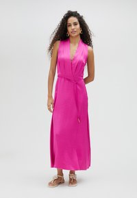 Vestido maxi de satén rosa con un profundo escote en V, diseño sin mangas, cintura ceñida con cinturón y aberturas laterales, combinado con sandalias de un color claro.