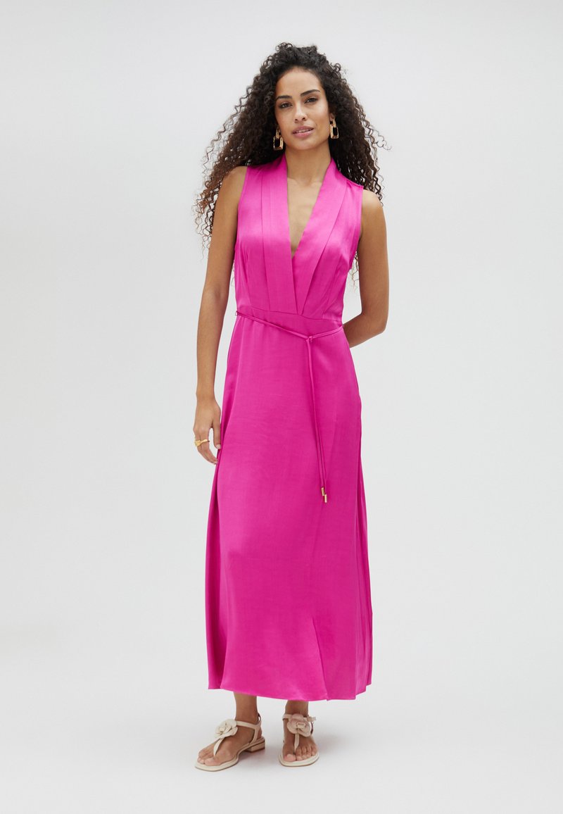 Vestido maxi de satén rosa con un profundo escote en V, diseño sin mangas, cintura ceñida con cinturón y aberturas laterales, combinado con sandalias de un color claro.