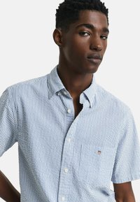 Kurzärmeliges Hemd mit Knopfleiste in hellblau und weißen vertikalen Streifen, mit einer einzelnen Brusttasche und kleinem Markenlogo auf der Tasche.