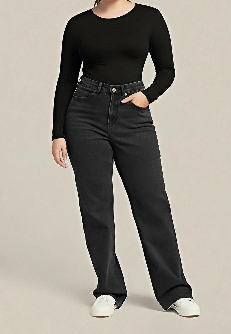 Femme portant un haut noir à manches longues et un jean noir taille haute, une main dans la poche, debout devant un fond uni.
