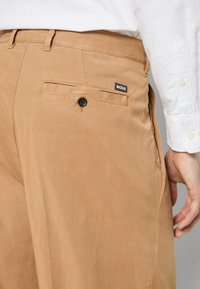 Μπεζ βαμβακερό παντελόνι με πίσω τσέπη, μαύρο κουμπί και μια μικρή μαύρη ετικέτα "BOSS" ραμμένη πάνω από την τσέπη. Λείο υλικό.