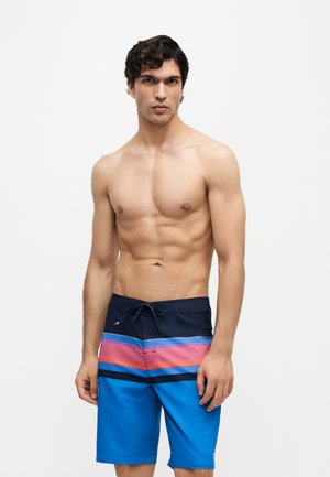 Ung mand med kort, krøllet hår iført blå, pink og sorte stribede badeshorts, står mod en ensfarvet hvid baggrund.
