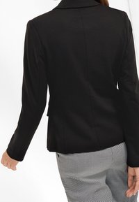 Personne portant un blazer noir ajusté et un pantalon à motif pied-de-poule noir et blanc, vue de dos avec les bras légèrement étendus.