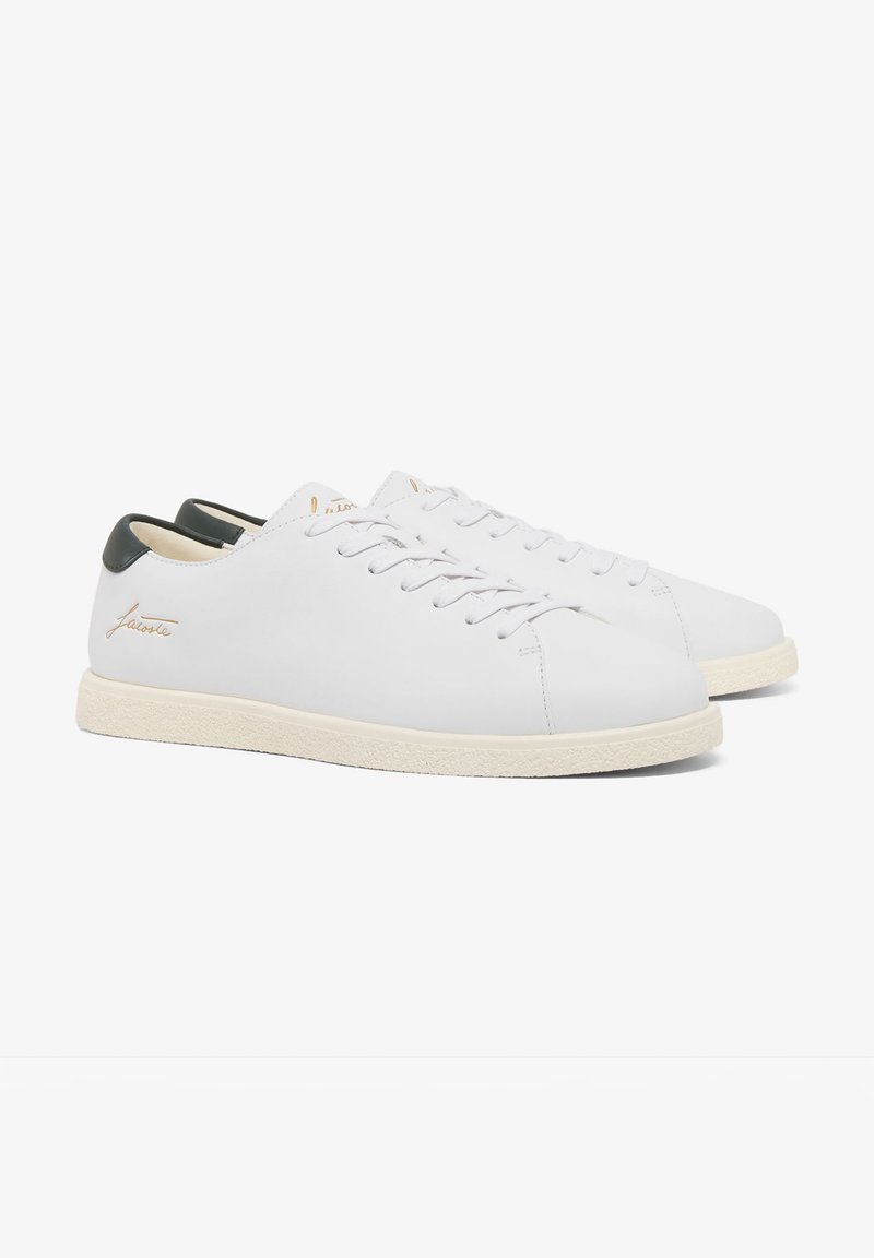 Sneakers basse in pelle bianca con suole color crema, linguette posteriori nere e scritta dorata del marchio sul lato esterno, mostrate fianco a fianco.